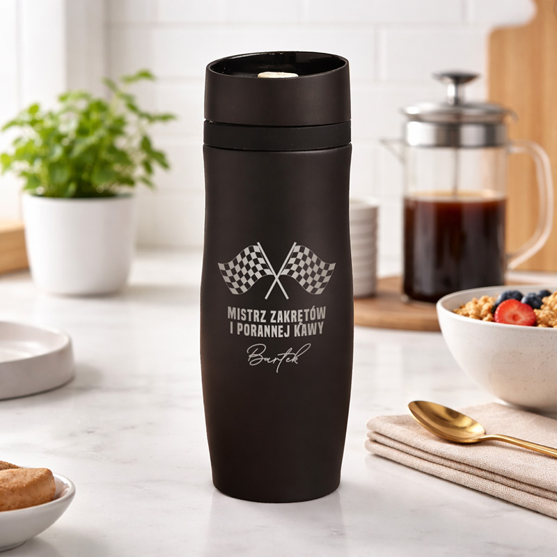 Czarny termos z logo "MISTRZ ZAKRĘTÓW I PORANNEJ KAWY" oraz flagami wyścigowymi, na tle kuchni z filiżanką kawy i płatkami.