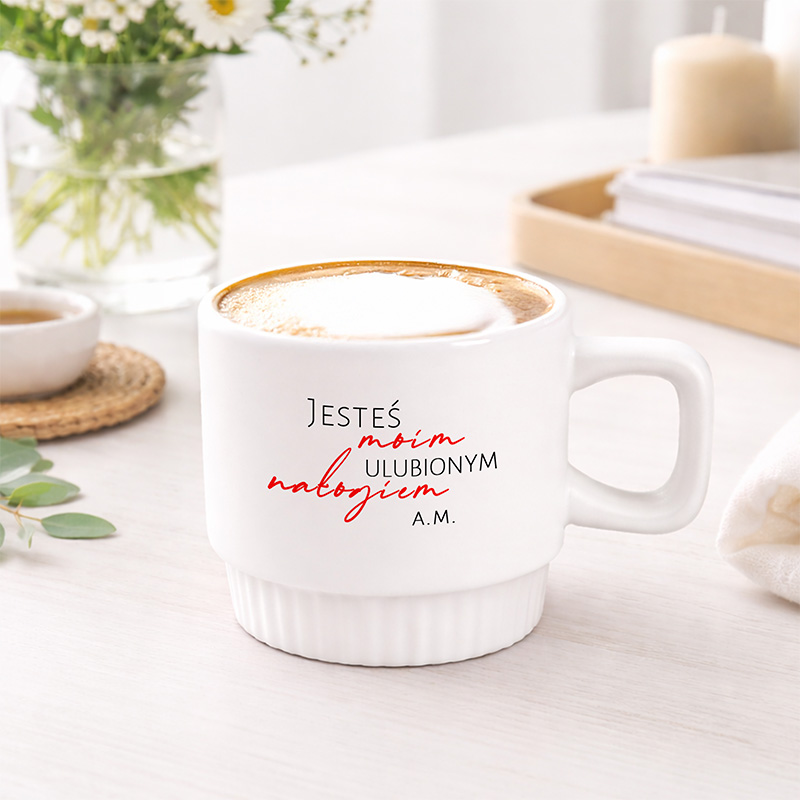 Biały kubek z napisem "Jesteś moim ulubionym nałogiem A.M." na brzegach, stojący na jasnym stole obok filiżanki i świecy.