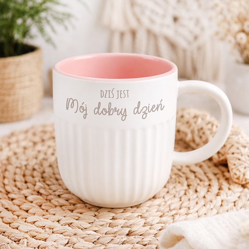 Kubek ceramiczny z różowym wnętrzem, z napisem "Dziś jest Mój dobry dzień" na białym tle. Otoczenie w stylu skandynawskim.