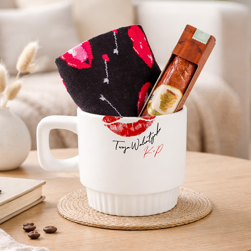 Kubek z napisem, wypełniony kolorową skarpetką i przekąską, stoi na okrągłym podkładce z juty. W tle widoczny stół i elementy dekoracyjne.