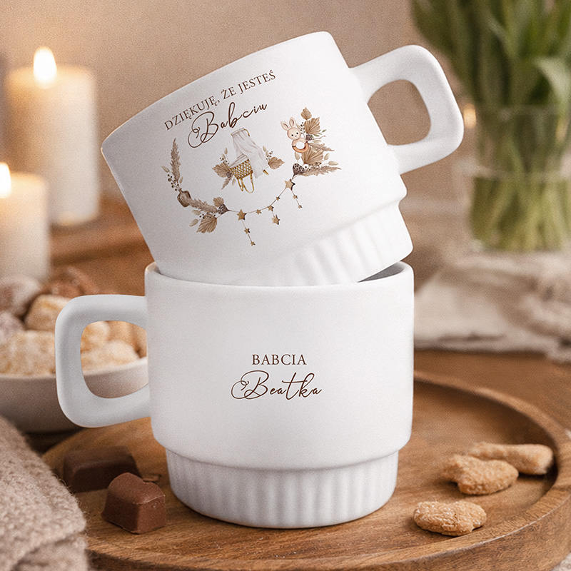 Dwie białe filiżanki, jedna na drugiej, z napisem "Dziękuję, że jesteś Babcią" oraz "Babcia Bełtka", w przytulnym otoczeniu.