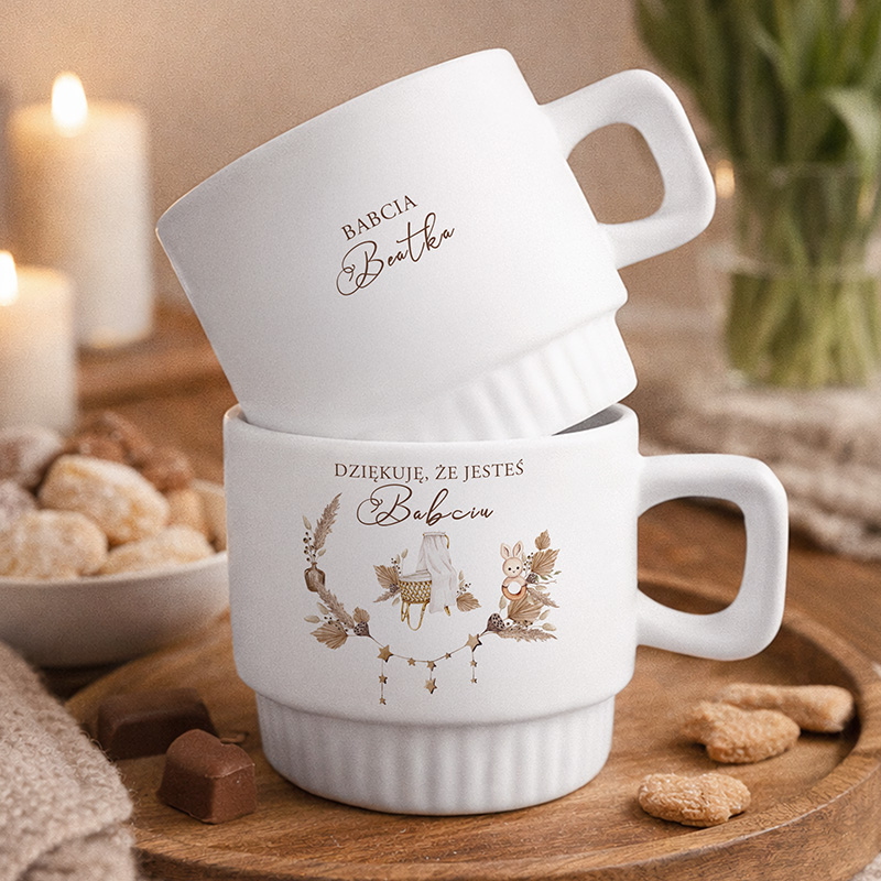 Dwie białe filiżanki ze złotymi napisami: "Babcia" i "Dziękuję, że jesteś Babciu", w otoczeniu świec i słodyczy.