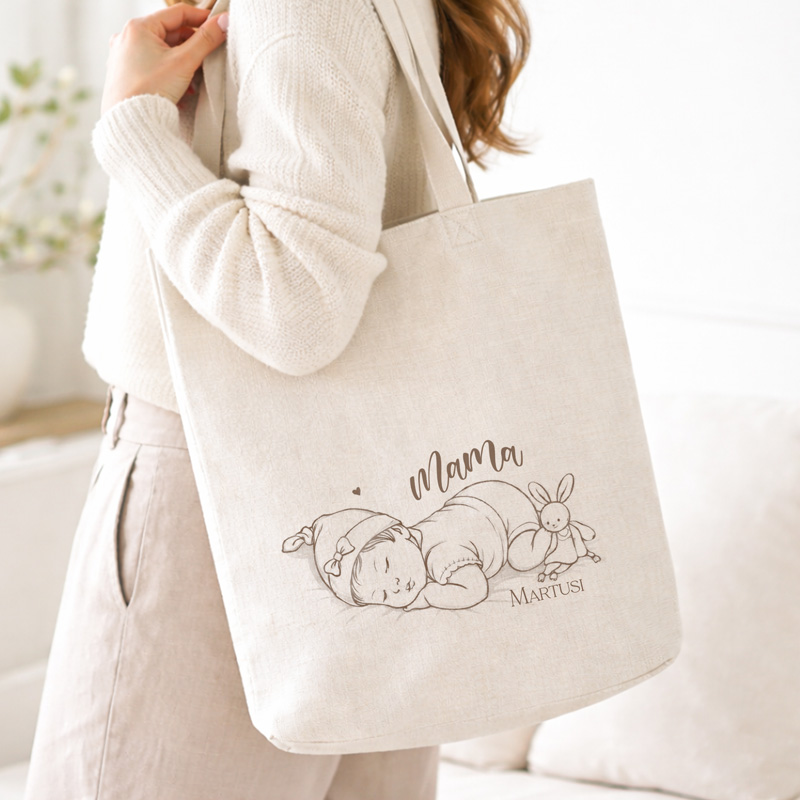 Torba materiałowa z rysunkiem śpiącego niemowlęcia, napisem "Mama" i króliczkiem, noszona przez osobę w jasnym swetrze.