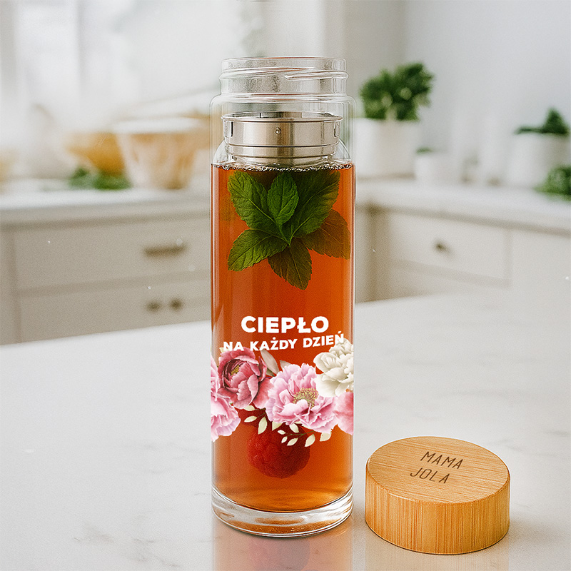 Przezroczysta butelka z herbatą, z widocznymi liśćmi mięty i kwiatami, na etykiecie napis "CIEPŁO NA KAŻDY DZIEŃ".