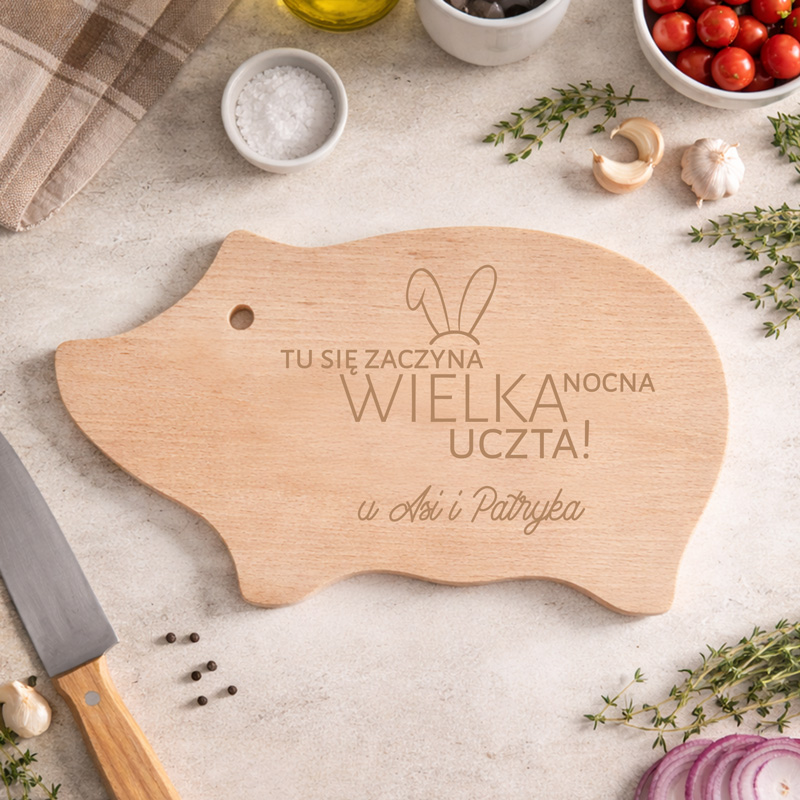 Deska do krojenia w kształcie świni z napisem "TU SIĘ ZACZYNA WIELKA NOCNA UCZTA!" oraz imionami", na kuchennym blacie z ziołami i przepisami.