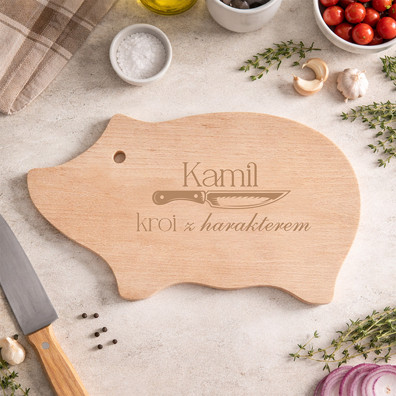 Deska do krojenia w kształcie świnki z napisem "Kamil kroi z harakterem" oraz nożem, otoczona ziołami i składnikami kuchennymi.