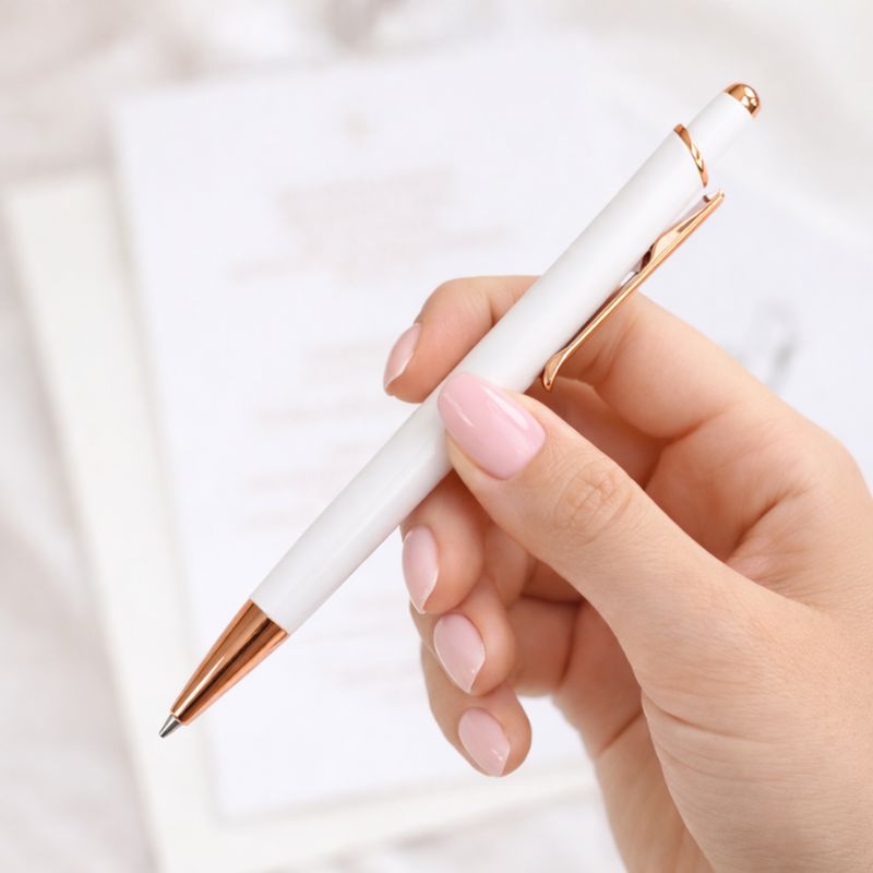 Biały długopis w ręce z różowymi paznokciami, na tle jasnych notatek.