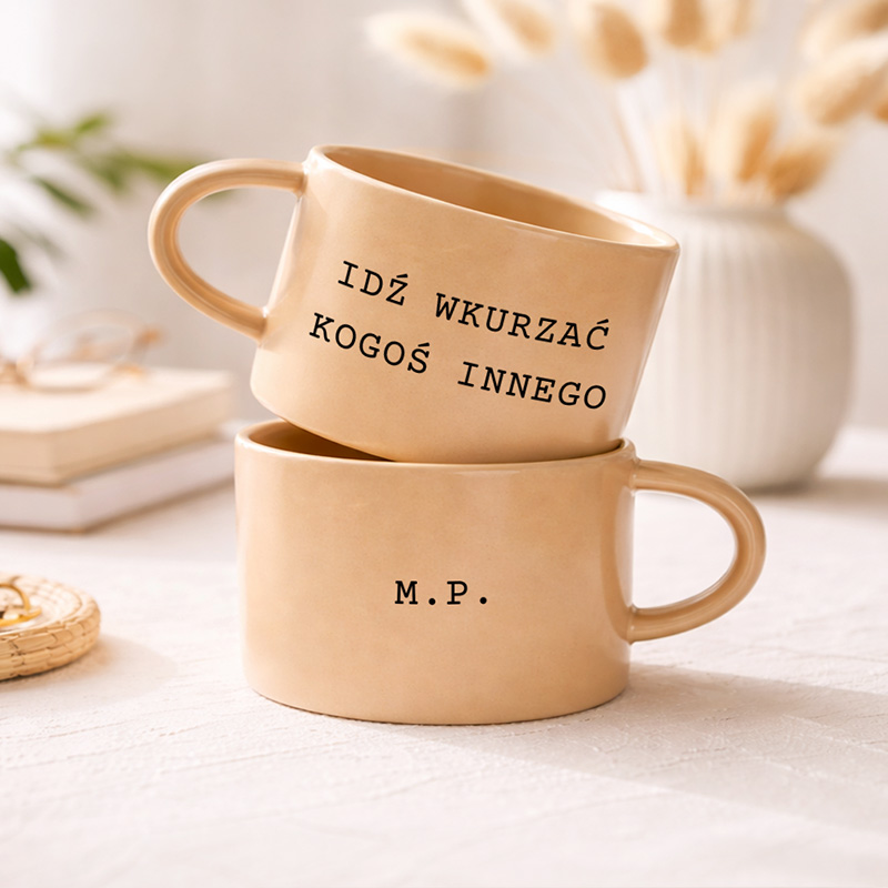 Dwa kubki w jasnym kolorze, jeden na drugim, z napisami "IDŹ WKURZAĆ KOGOŚ INNEGO" i "M.P."