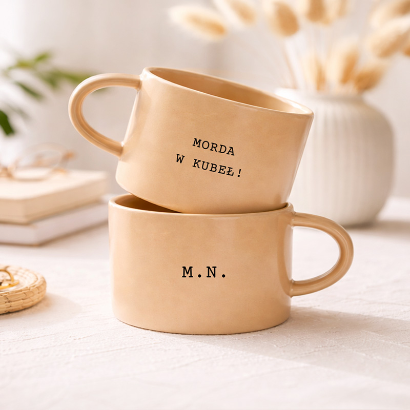 Dwie filiżanki w kolorze beżowym, jedna na drugiej, z napisem "MORDA W KUBEL!" na górnej i "M.N." na dolnej.