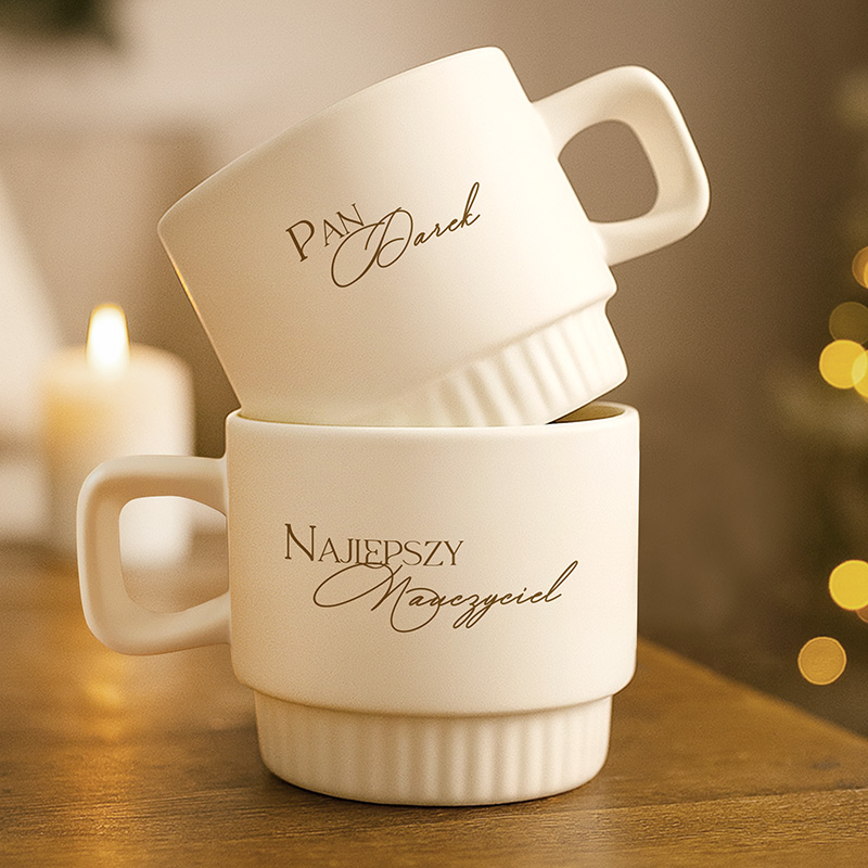 Dwa kremowe kubki z dekoracyjnymi napisami: "Pan IMIĘ" i "Najlepszy Nauczyciel". Tło z miękkim oświetleniem.