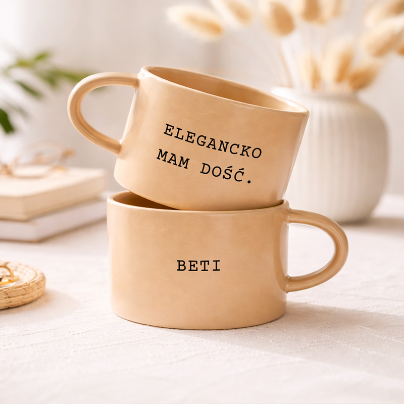 Dwa kubki na stole, jeden na drugim; jeden z napisem "ELEGANCKO MAM DOŚĆ." i drugi z imieniem "BETI".