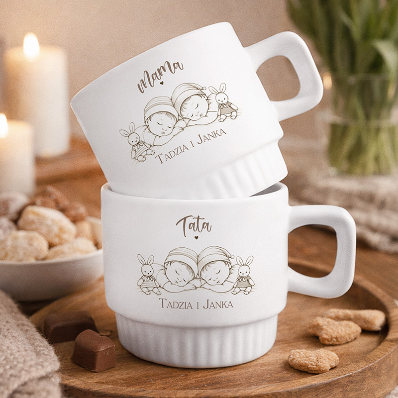 Dwie białe filiżanki z rysunkami dzieci i napisami "Mama" oraz "Tata", stojące na drewnianym stole obok słodkości.