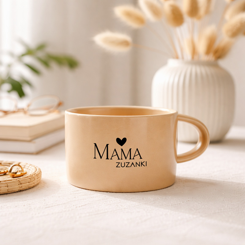 Kremowy kubek z napisem "MAMA" i imieniem, stojący na stole obok dekoracyjnego koszyczka z biżuterią.