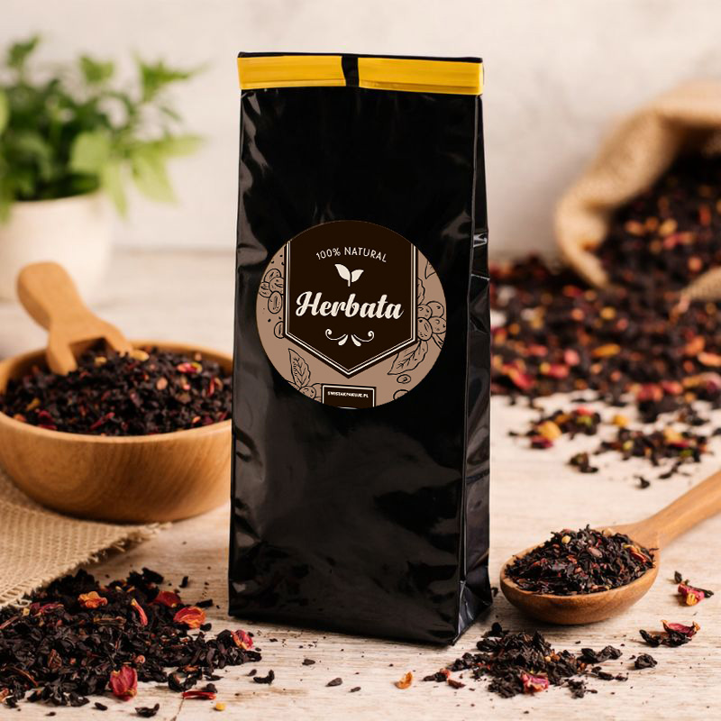 Czarna torebka z herbatą, z etykietą "100% Natural", otoczona suszonymi herbata i drewnianą łyżką.