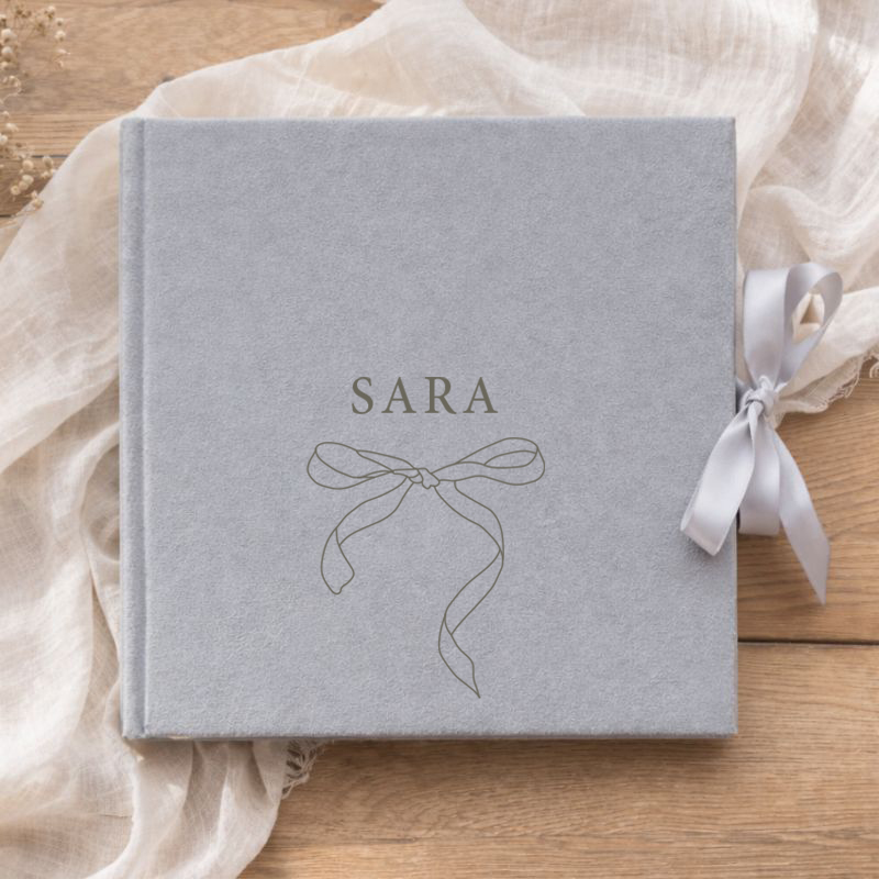 Szara okładka albumu z imieniem i rysunkiem wstążki, leżącego na jasnym materiałowym tle.