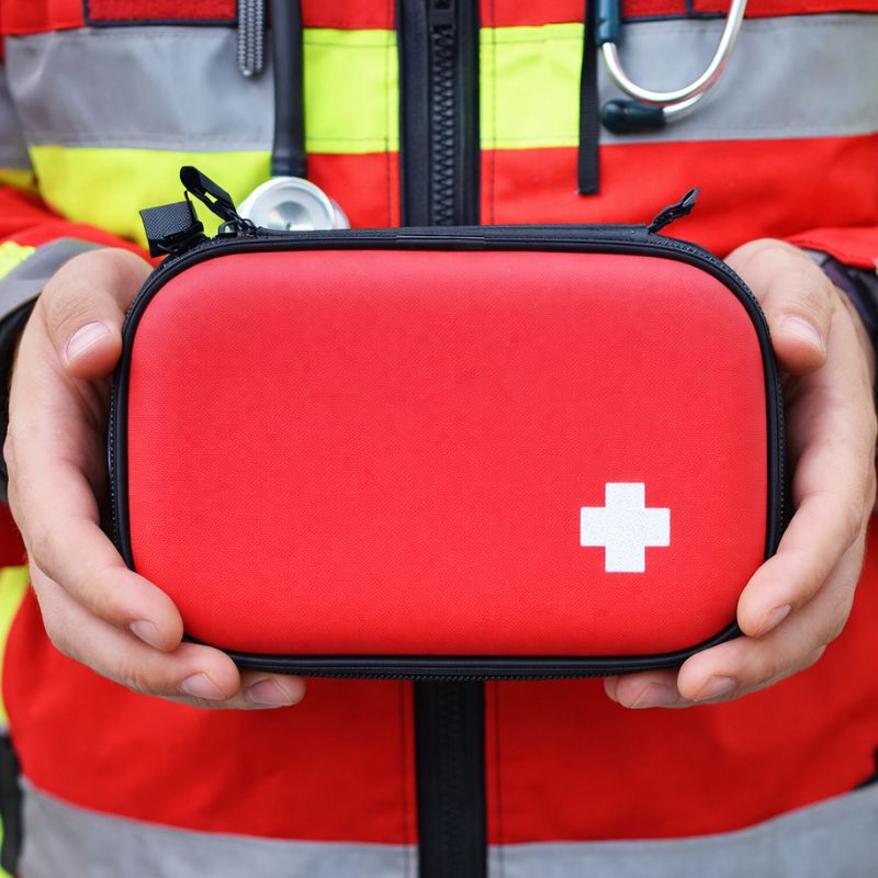 Czerwony plecak medyczny z białym krzyżem, trzymany przez osobę w odzieży ratunkowej.