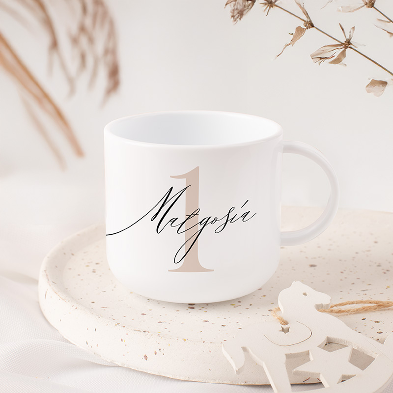 Biała filiżanka z imieniem i dużą cyfrą "1", ustawiona na jasnej, okrągłej podstawce. W tle suszone kwiaty.