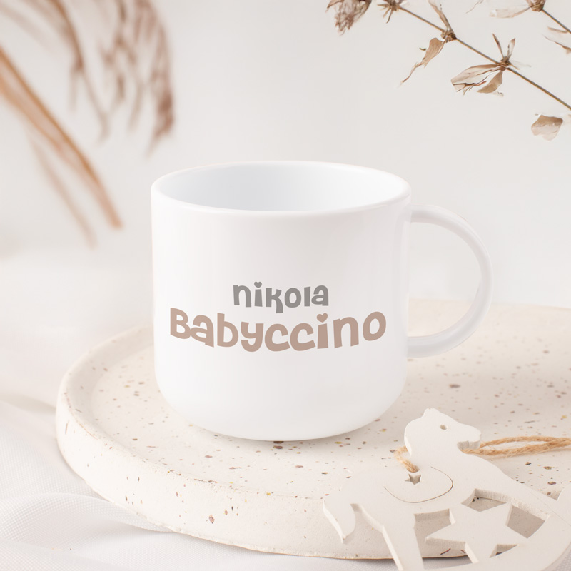 Biała filiżanka z napisem „Babyccino” na gładkiej, jasnej powierzchni w aranżacji z dodatkami dekoracyjnymi.