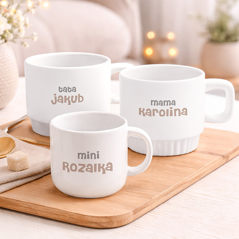 Trzy białe filiżanki na drewnianej desce, z napisami "tata Jakub", "mama Karolina" i "mini Rożańka".