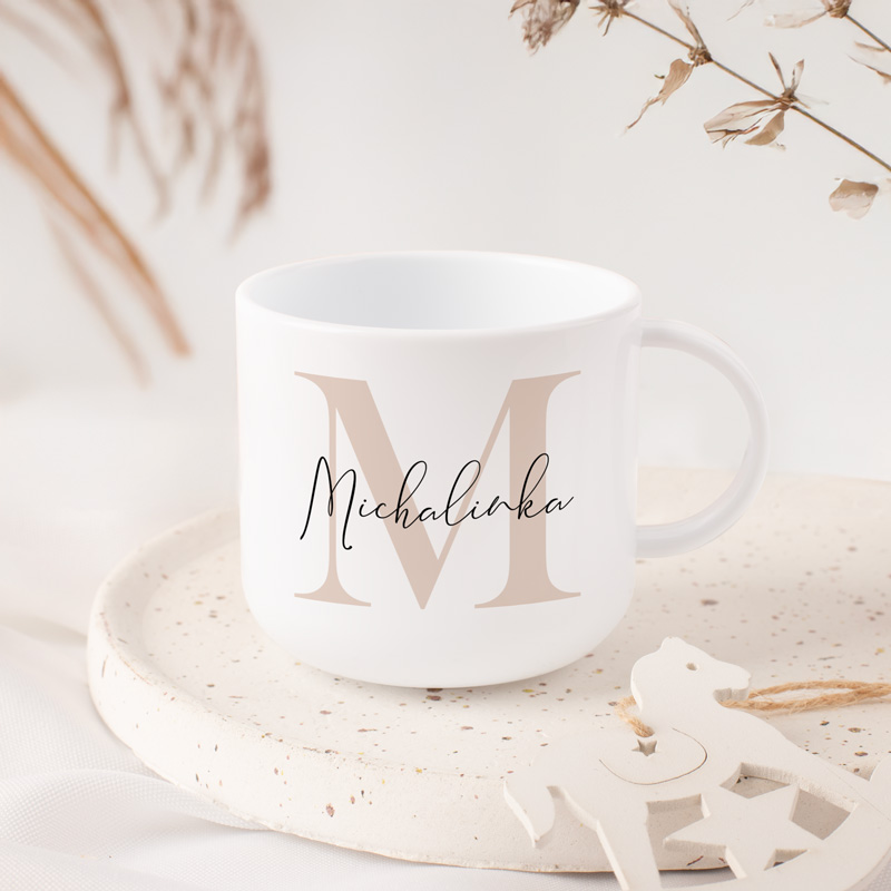 Biała filiżanka z dużą literą oraz imieniem na powierzchni. W tle delikatne dekoracje roślinne.