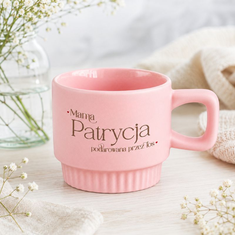 Różowy kubek z napisem "Mama Patrycja podarowana przez lós", ustawiony na drewnianym stole, w otoczeniu kwiatów.