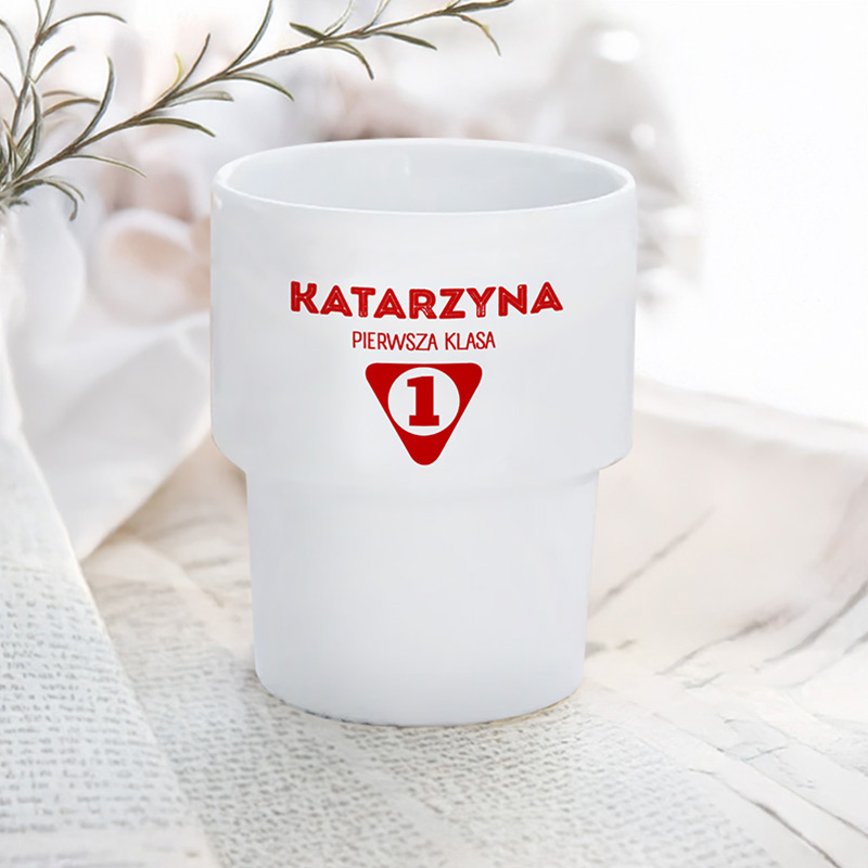 Biały kubek z czerwonym napisem "KATARZYNA" oraz oznaczeniem "PIERWSZA KLASA" w czerwonym trójkącie z numerem "1".