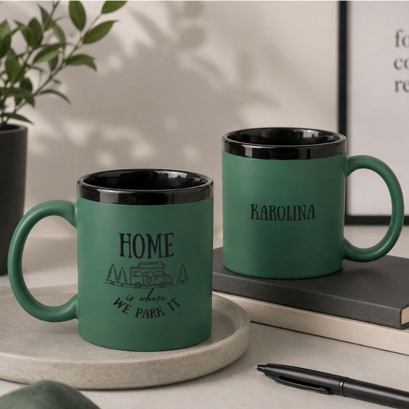 Zielone kubki ceramiczne na szarej tacy, jeden z napisem "HOME is where we park it", drugi z imieniem "KAROLINA". W tle rośliny.