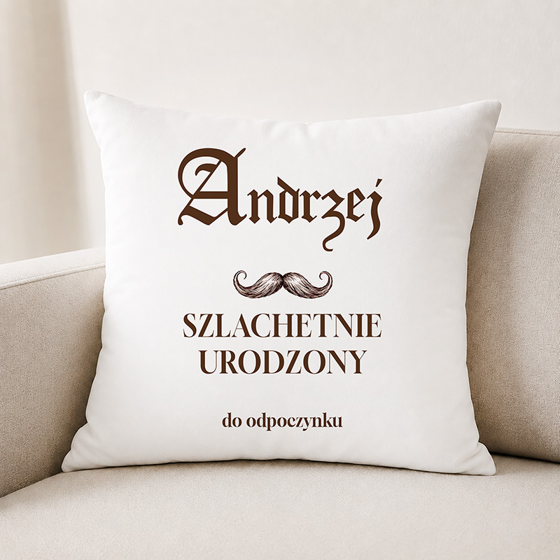 Poduszka z napisem "Andrzej Szlachetnie Urodzony" oraz ozdobnym wąsem, leżąca na jasnej sofie.